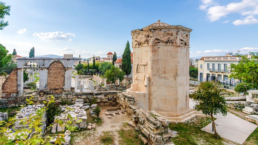 Tower of Winds in Roman Agora, Athens, with ancient ruins and cityscape in the background.
