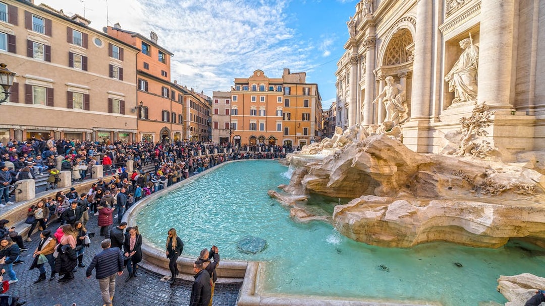 Crowd gathered around Trevi Fountain in Rome, Italy, with historic architecture in the background.