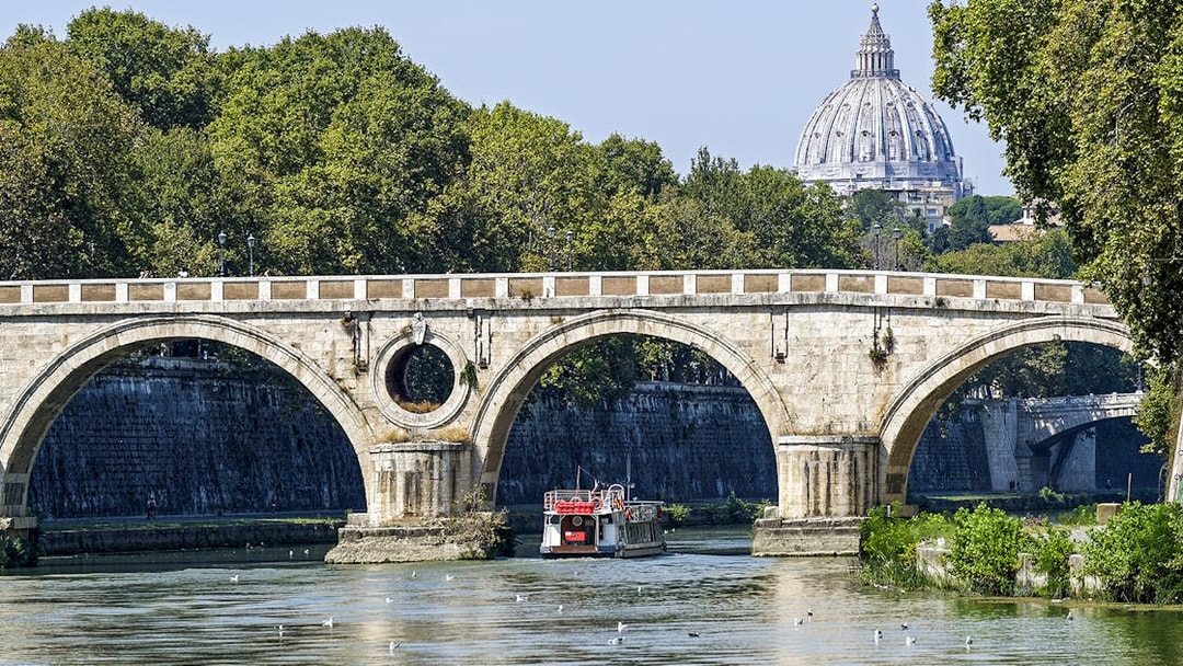 Hop-on hop-off river cruise boat under Rome's Ponte Sant'Angelo with St. Peter's Basilica in view.