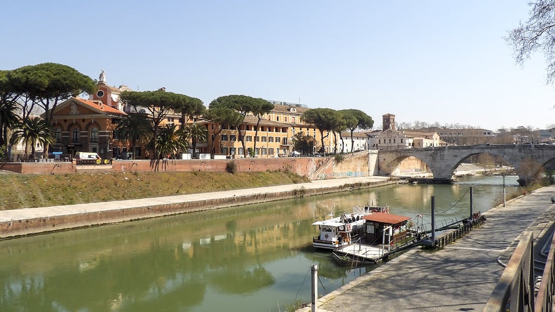 Hop-on hop-off river cruise boat docked on the Tiber River in Rome, near historic buildings and a stone bridge.