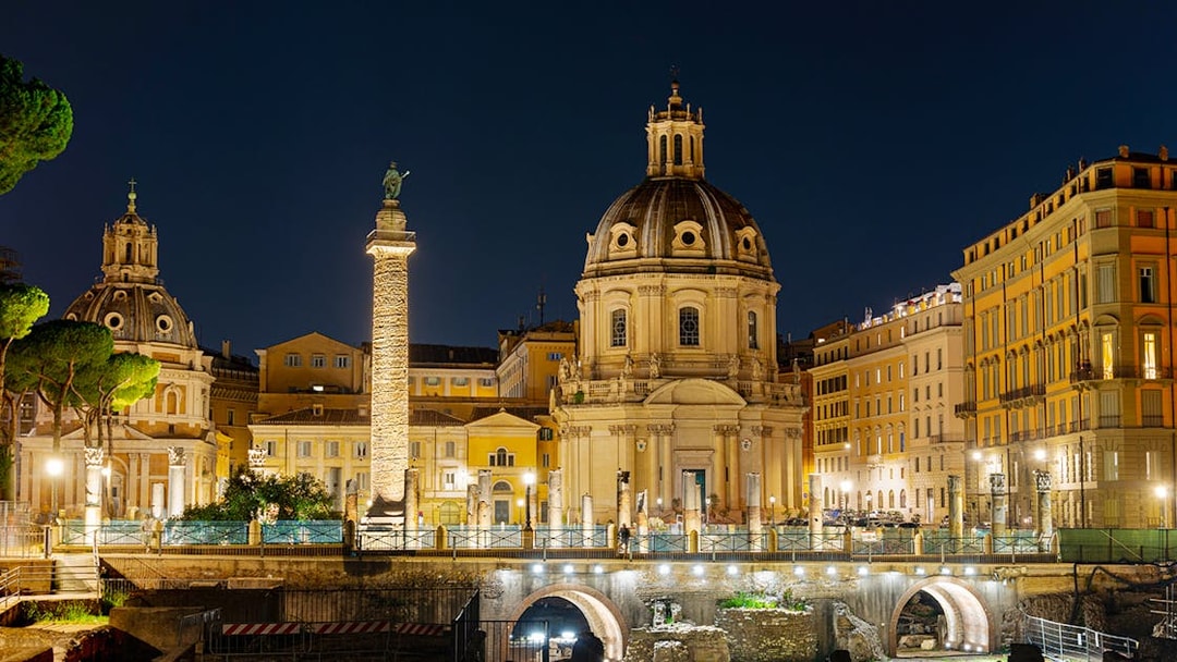 Fori Imperiali illuminated at night with Trajan's Column and surrounding architecture in Rome.