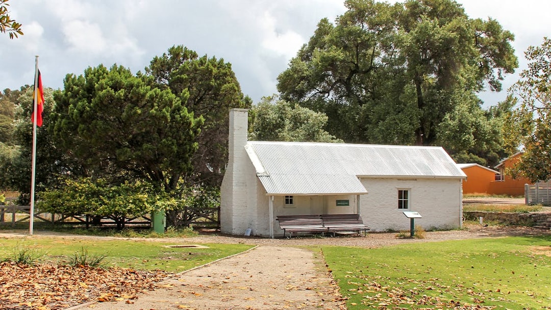 Historic cottage on Rottnest Island surrounded by trees, with a flagpole and pathway in view.