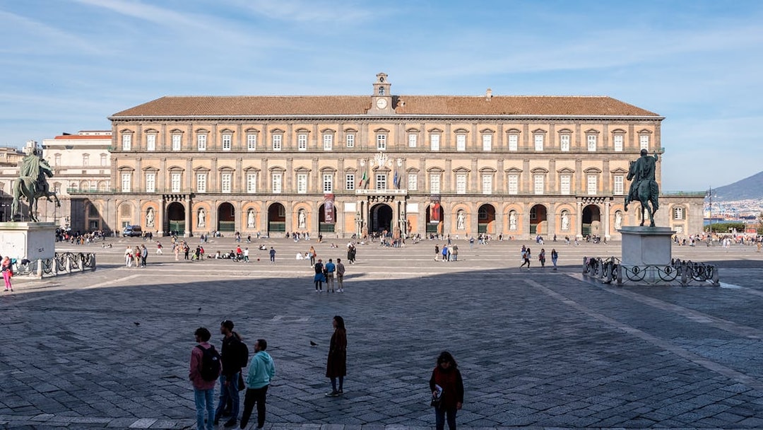 Royal Palace of Naples facade with equestrian statues and visitors in the foreground.