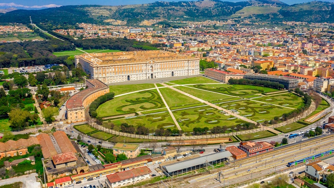 Aerial view of the Royal Palace of Caserta in Italy with expansive gardens and surrounding cityscape.