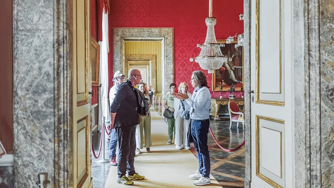 Tour group with guide inside Royal Palace of Caserta, ornate red room with chandelier.