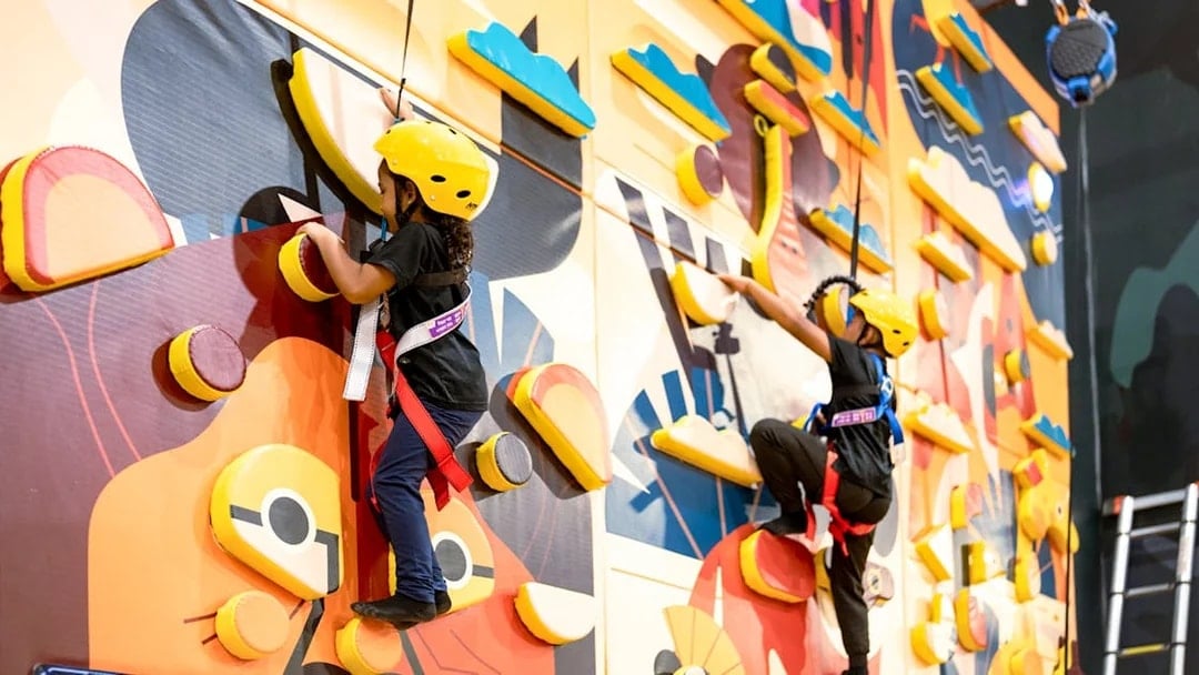 Children climbing a colorful indoor rock wall with safety harnesses.