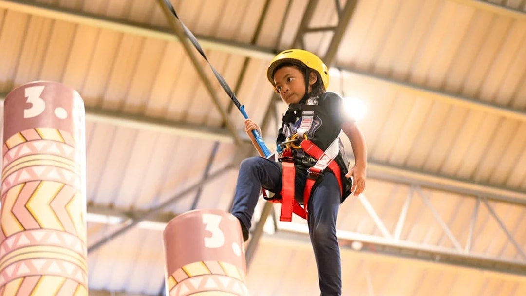 Child balancing on a pole in an indoor activity center, wearing a safety harness and helmet.
