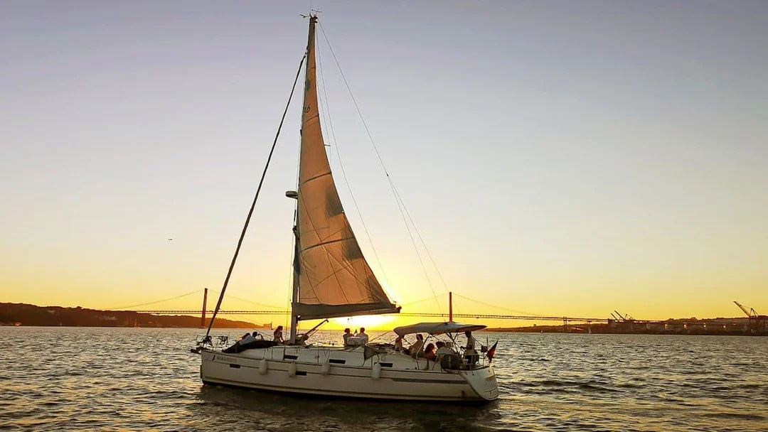 Sailboat on Tagus River at sunset with Lisbon's 25 de Abril Bridge in the background.