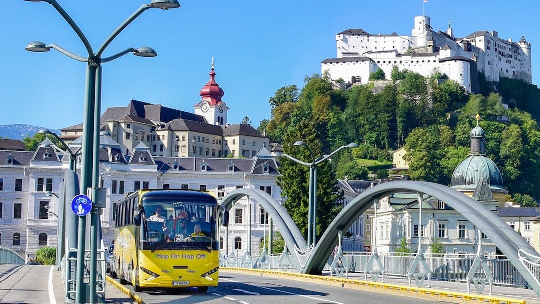 Hop On Hop Off bus crossing a bridge in Salzburg with Hohensalzburg Fortress in the background.