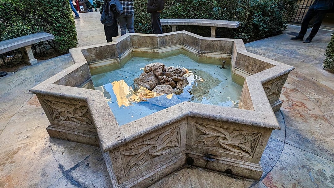 Stone fountain in the courtyard of San Nicolas Church, surrounded by benches and greenery.