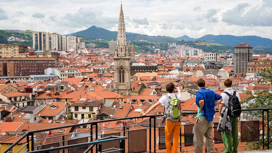 Tourists overlooking Bilbao cityscape with view of church spire and surrounding hills.
