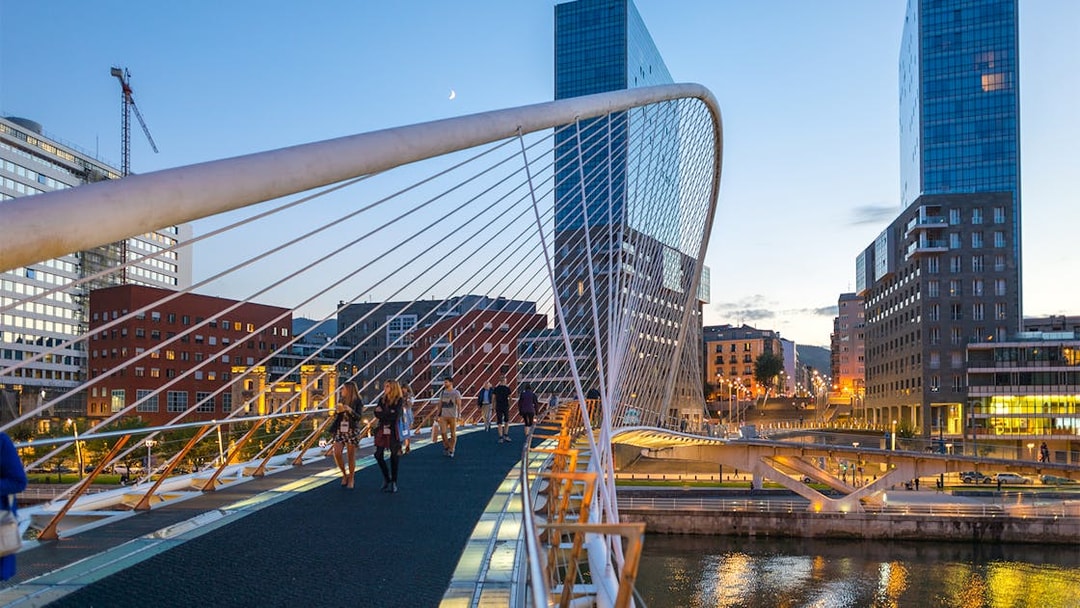 Pedestrians on Zubizuri Bridge in Bilbao at dusk, with cityscape and river views.
