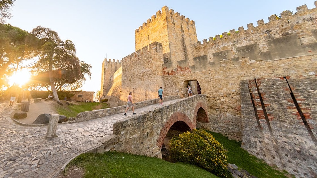 Visitors walking across a stone bridge towards St. George’s Castle entrance in Lisbon.