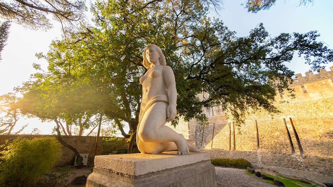 Statue of a woman at Sao Jorge Castle, Lisbon, with trees and castle wall in the background.
