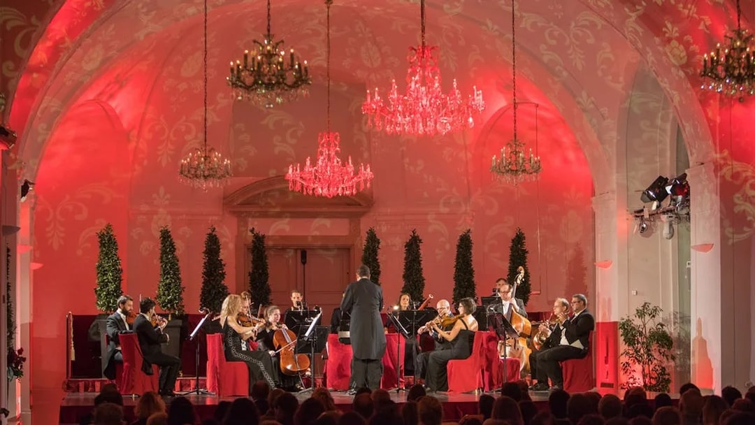 Orchestra performing at Schönbrunn Palace concert hall with chandeliers and red lighting.