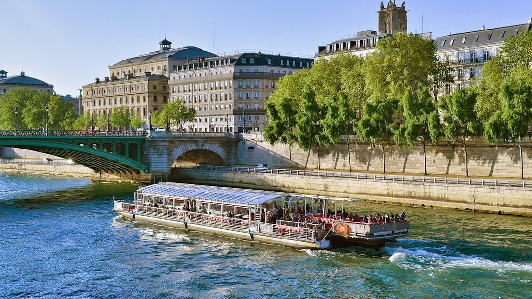 Seine River cruise boat passing under a bridge in Paris with historic buildings in view.