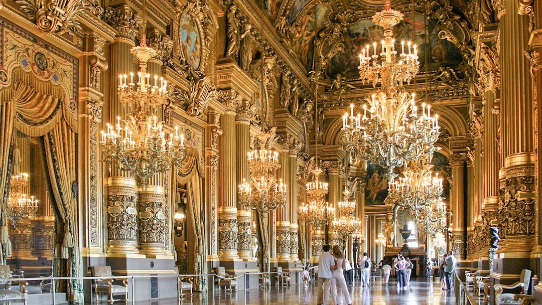 Visitors inside Opéra Garnier, Paris, admiring ornate chandeliers and gilded architecture.