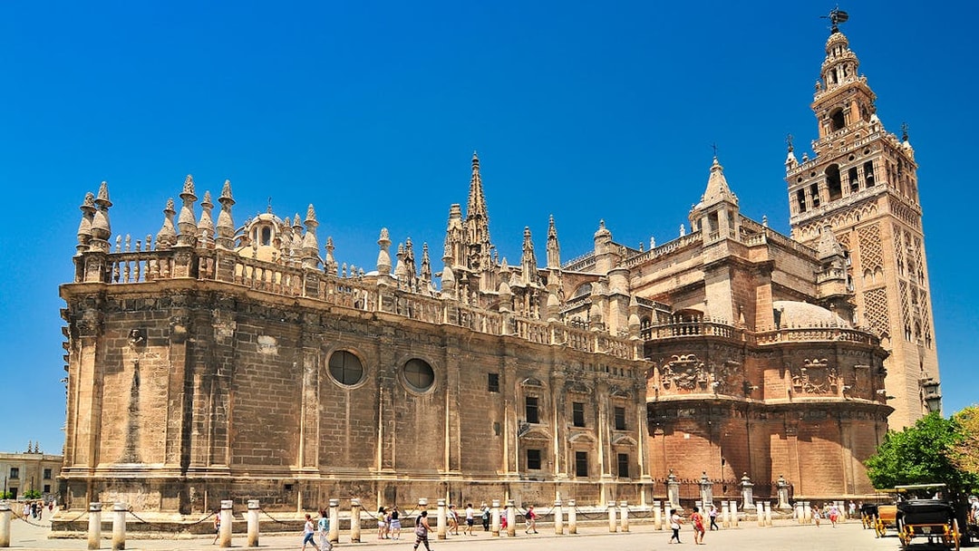 Seville Cathedral and Giralda tower under a clear blue sky, with tourists exploring the plaza.