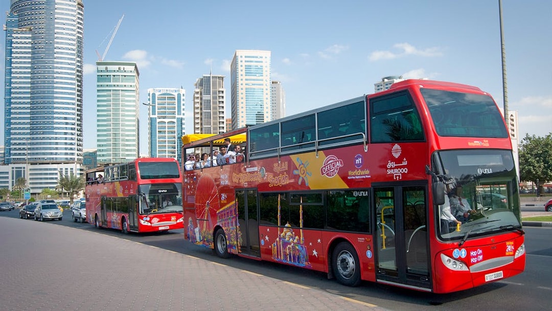 Red double-decker bus on Sharjah Hop-On-Hop-Off tour with city skyscrapers in the background.