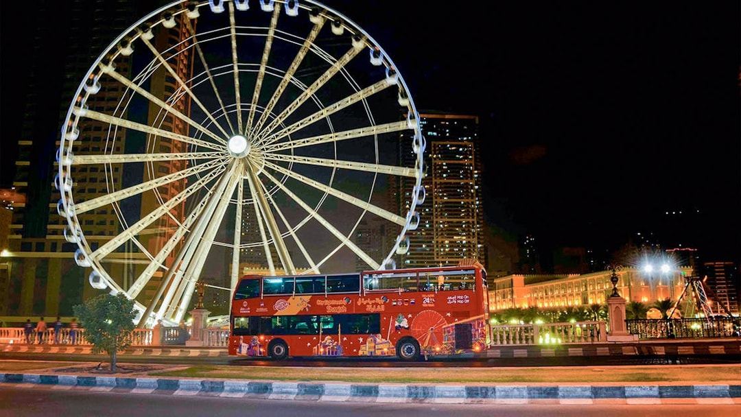 Red double-decker bus in front of illuminated Ferris wheel on Sharjah Hop-On-Hop-Off Bus Tour.