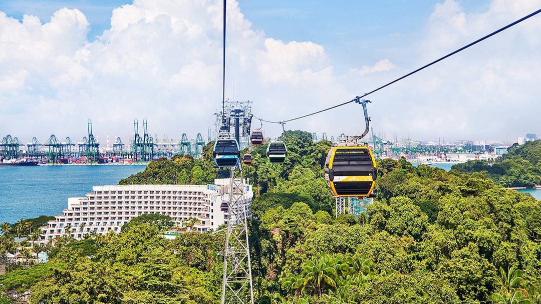 Cable cars over Sentosa Island with view of lush greenery and Singapore harbor.