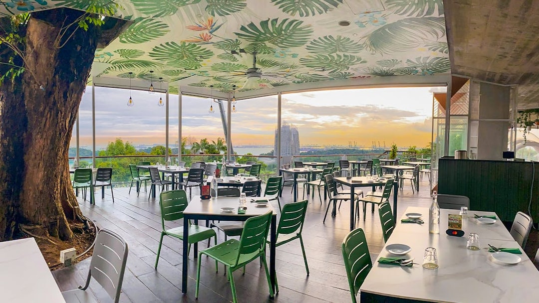 Outdoor dining area with view of Singapore skyline at sunset, featuring lush greenery and modern decor.