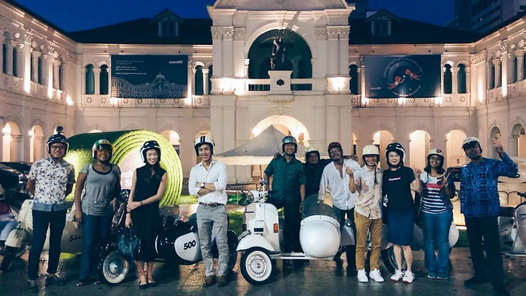 Group of people with vintage Vespas in front of Singapore Art Museum at night.