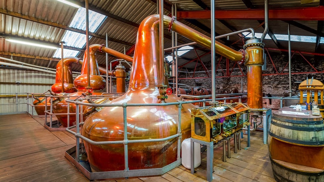 Copper stills inside a whiskey distillery with wooden barrels and equipment.
