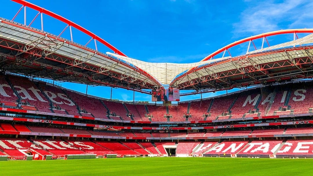 Benfica Stadium ground view with red seating and green field under blue sky.