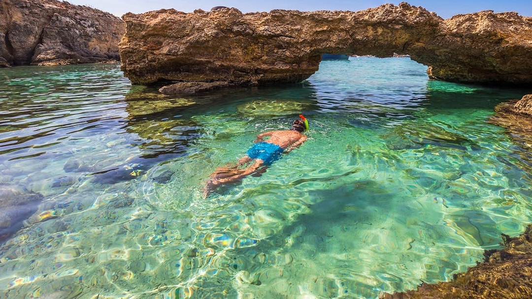 Snorkeler in clear waters near rock arch in Comino, Malta.