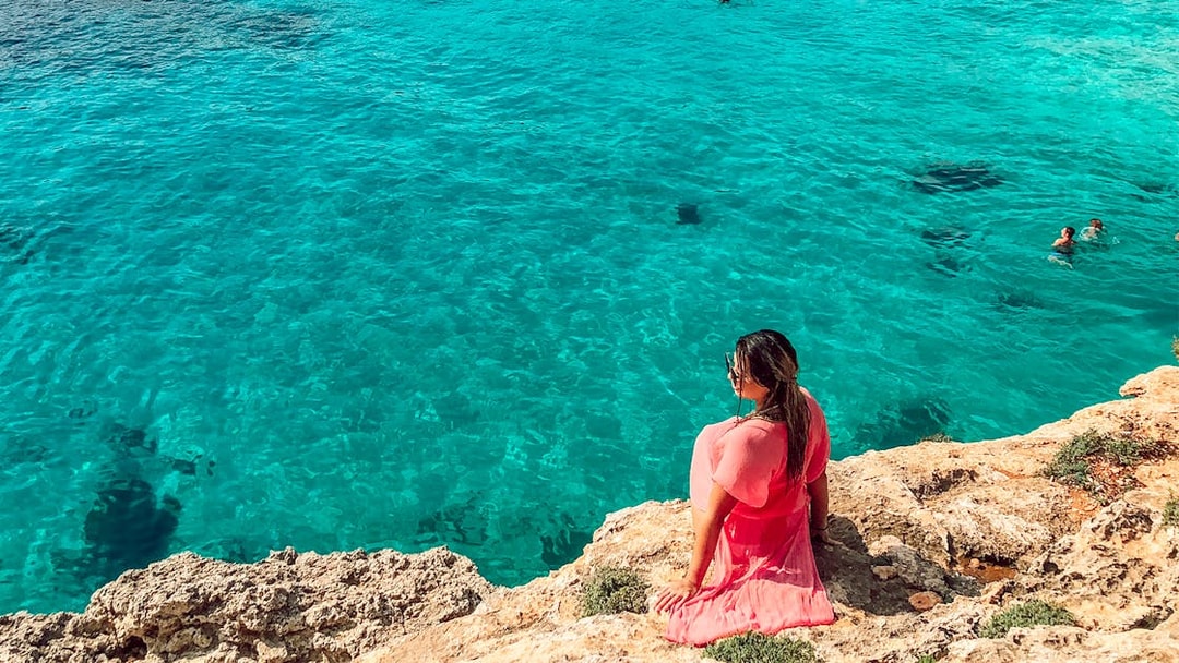 Woman in pink dress sitting on rocky shore overlooking clear blue water in Comino, Malta.