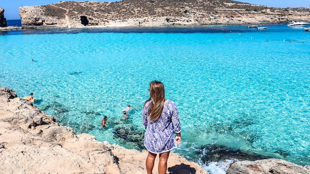 Woman overlooking clear blue waters at Comino, Malta, with swimmers and rocky coastline.