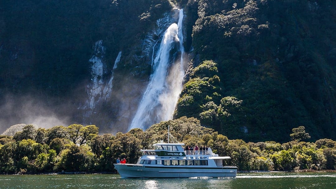 Small boat cruising near waterfall in Milford Sound, New Zealand.