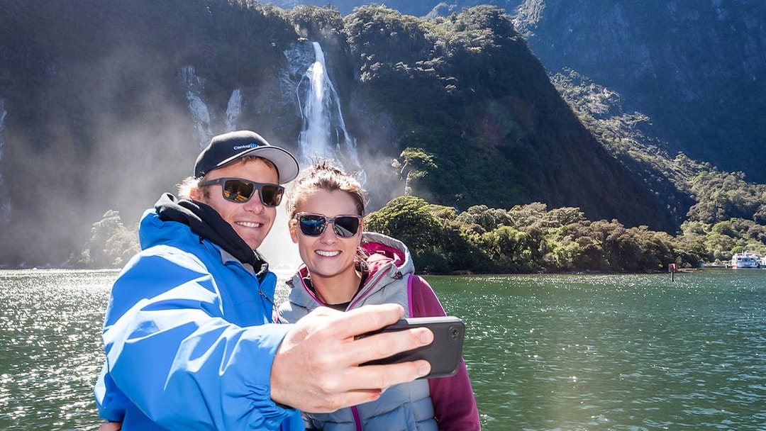 Couple taking selfie on Milford Sound cruise with waterfall in background.