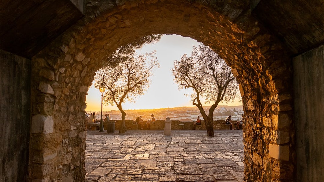 Archway view of sunset at St. George’s Castle, Lisbon, with people sitting near trees.