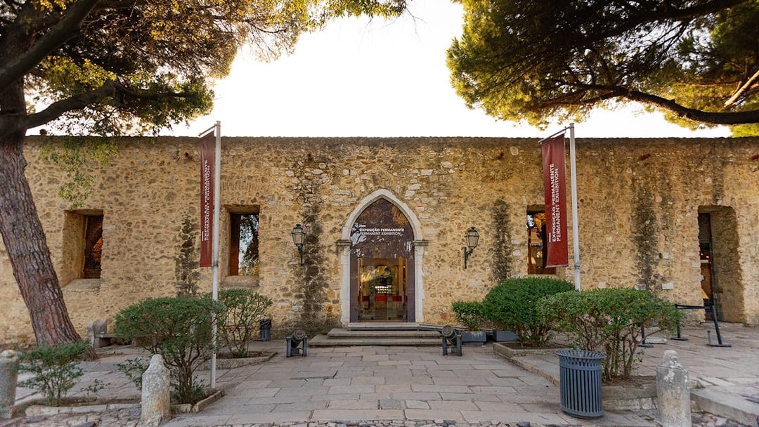 Entrance to Permanent Exhibitions at St George's Castle, Lisbon, with stone facade and flags.