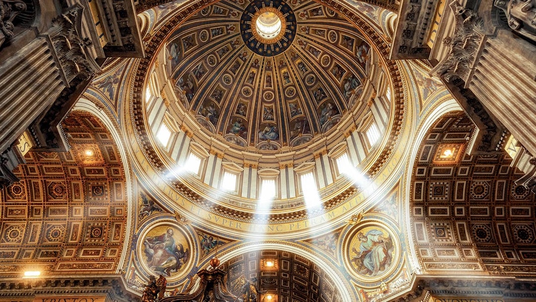 Dome and ornate ceiling of St. Peter’s Basilica with sunlight streaming through windows.