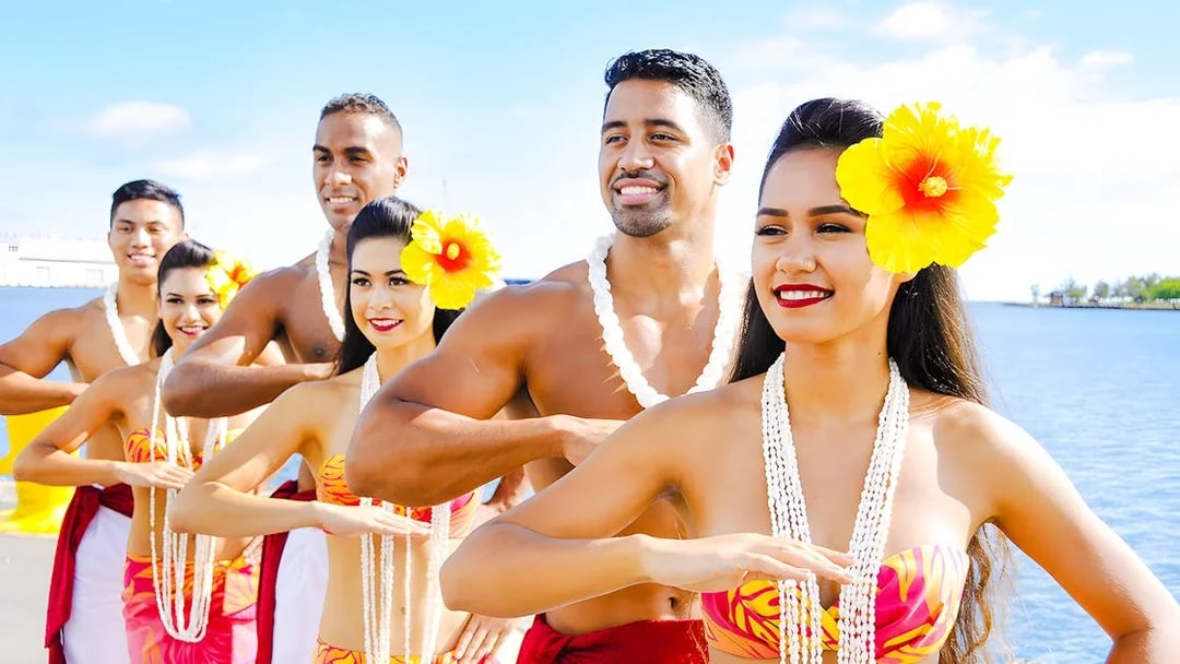 Hawaiian dancers in traditional attire with leis performing by the ocean.