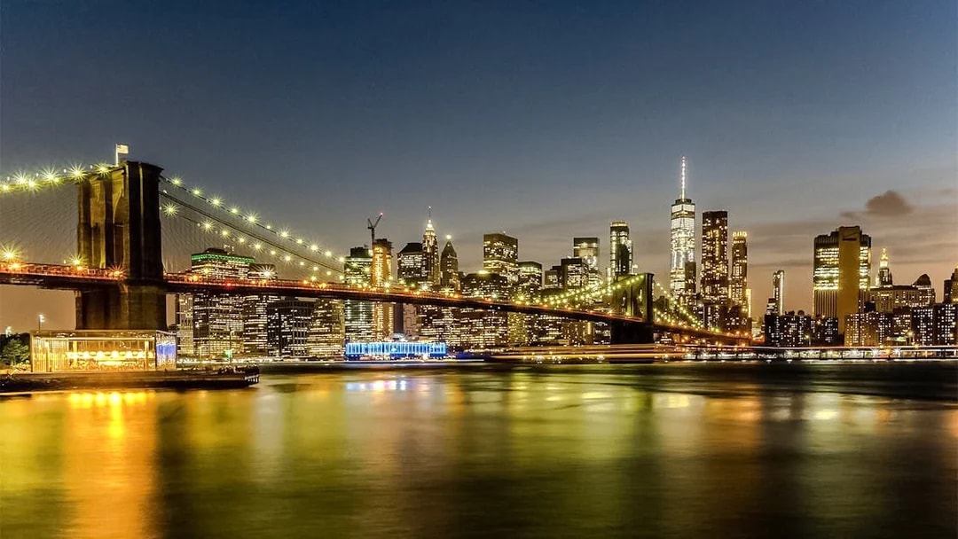 Brooklyn Bridge and Manhattan skyline illuminated at sunset during Statue of Liberty & Ellis Island cruise.