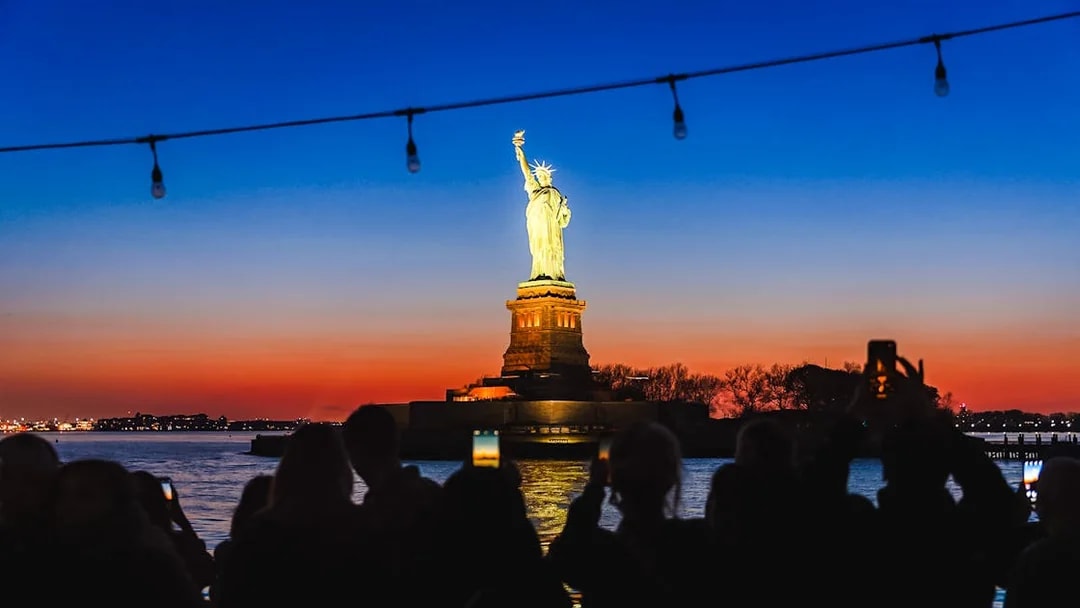 Statue of Liberty illuminated at night with sunset sky, viewed from a boat.