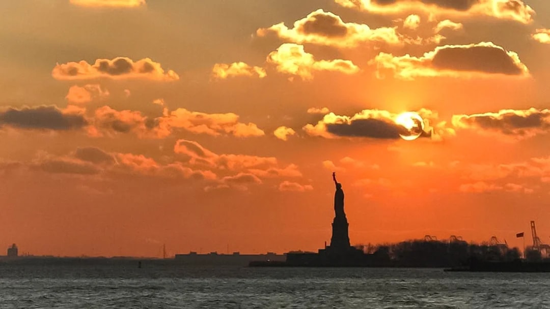 Statue of Liberty silhouetted against sunset sky during Ellis Island cruise.