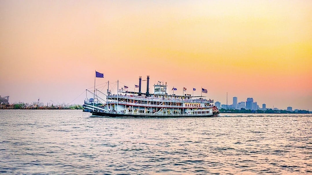 Steamboat Natchez cruising at sunset with city skyline in the background.