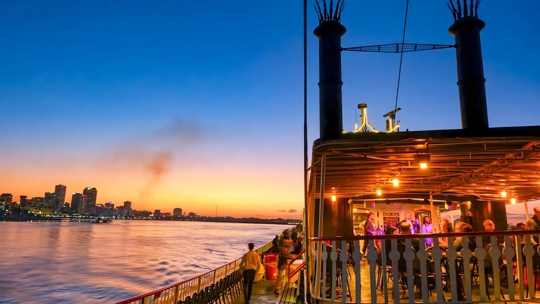 Steamboat Natchez cruising on the Mississippi River at sunset with New Orleans skyline.