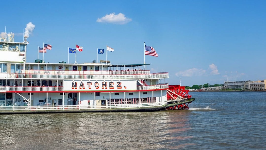 Steamboat Natchez departing from dock on a sunny day with flags flying.