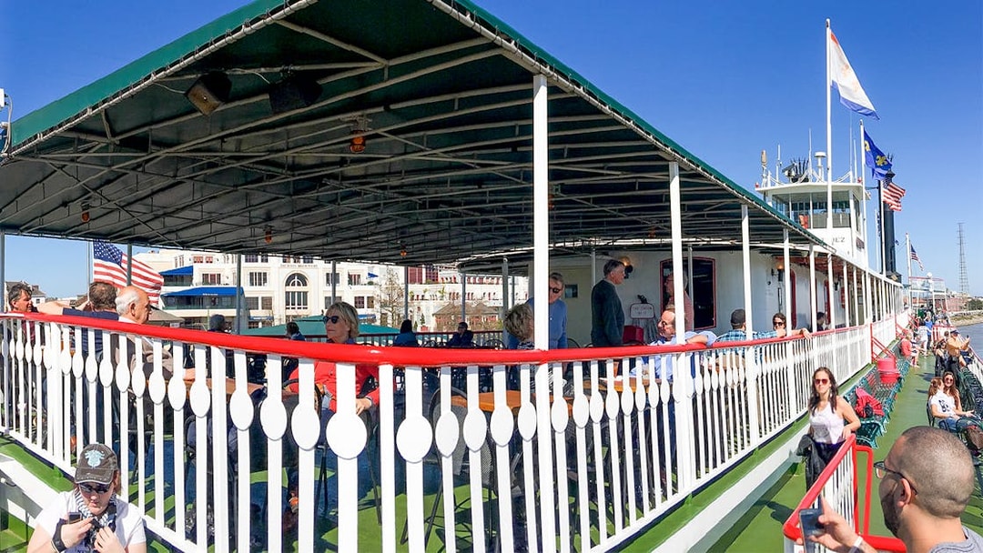 Steamboat Natchez deck with passengers, New Orleans skyline in background on Mississippi River.