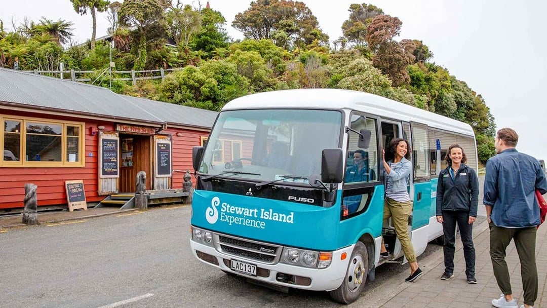 Stewart Island tour bus parked near a red building with people boarding and interacting.