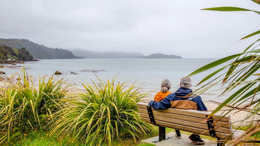 Couple sitting on a bench overlooking the beach and bay on Stewart Island tour.
