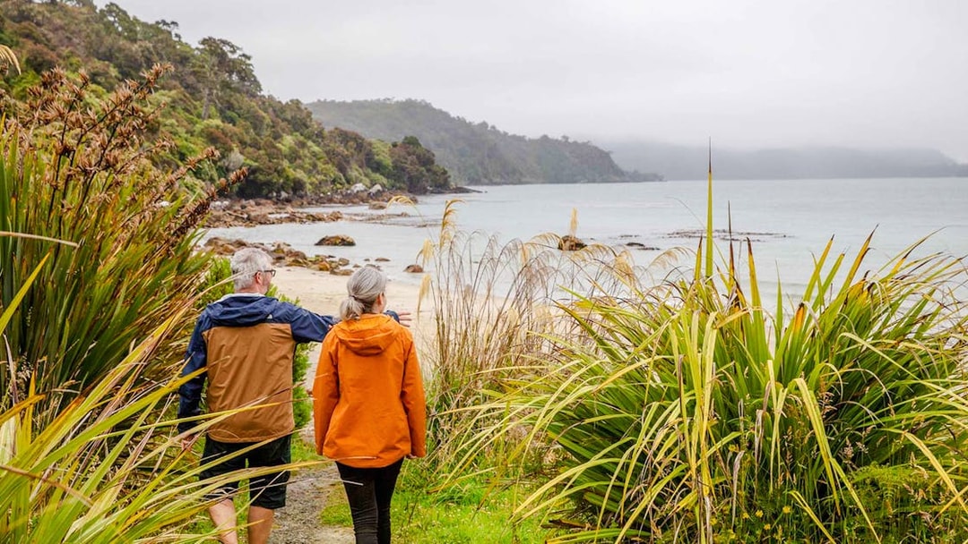 Couple walking along a coastal path on Stewart Island, surrounded by lush greenery and ocean views.
