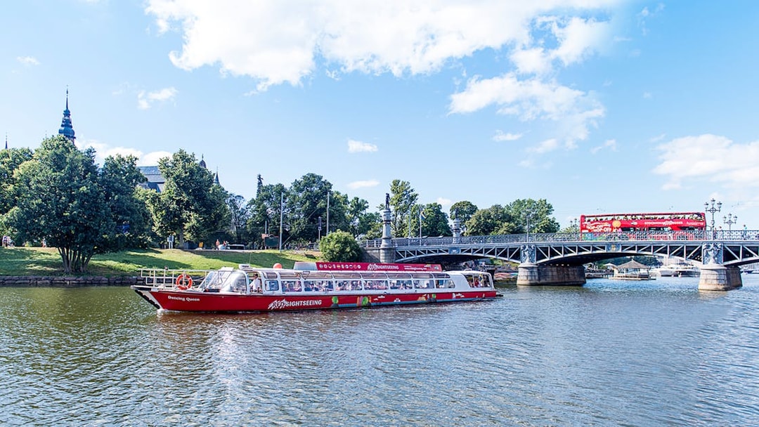 Red sightseeing boat on Stockholm's waterway near a bridge and greenery.