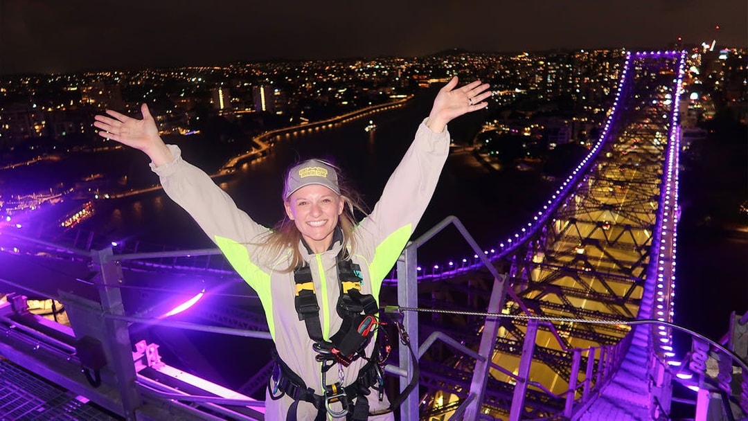 Person celebrating atop illuminated Story Bridge during night climb in Brisbane.